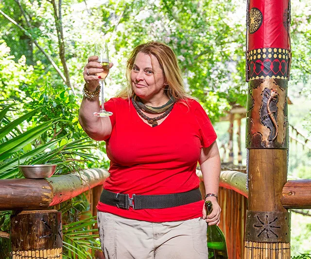 Person in red shirt holding a wine glass, standing on a wooden walkway in a lush jungle setting.