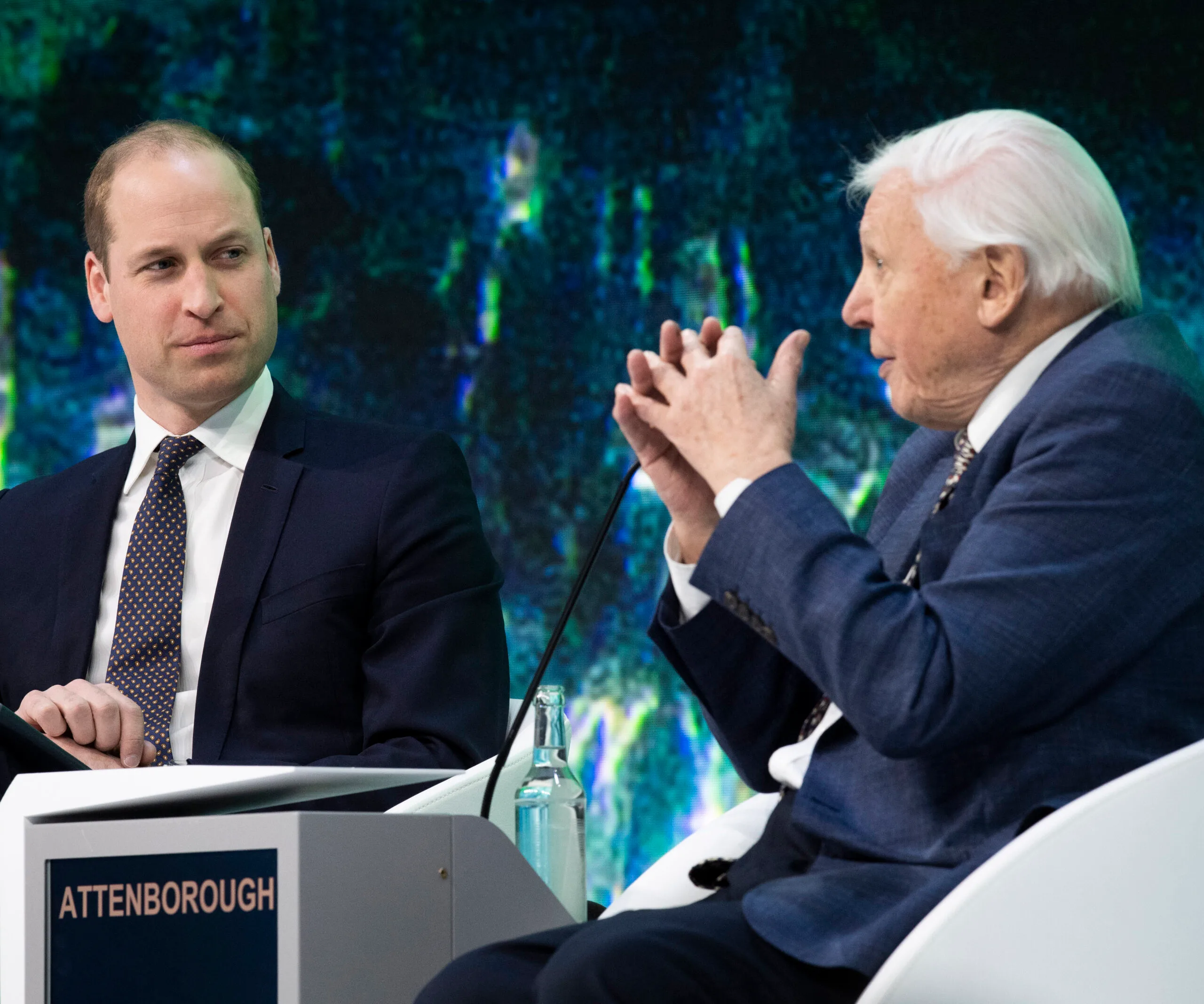 Two men in suits in conversation at an event, seated, with a podium labeled "ATTENBOROUGH."