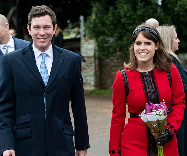 A smiling couple dressed elegantly, the woman holding a bouquet of flowers, walking outside on a cloudy day.