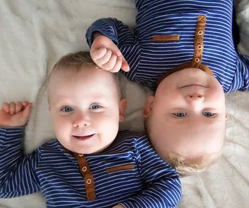 Twin babies lying on a bed, both wearing matching blue and brown striped shirts.