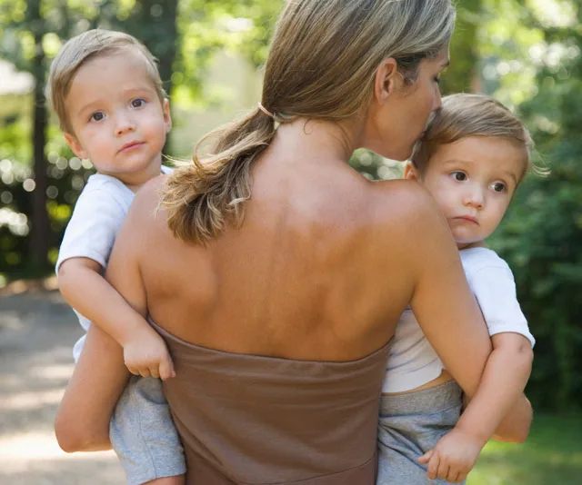 A woman carries two toddlers outdoors in a green, sunlit setting.