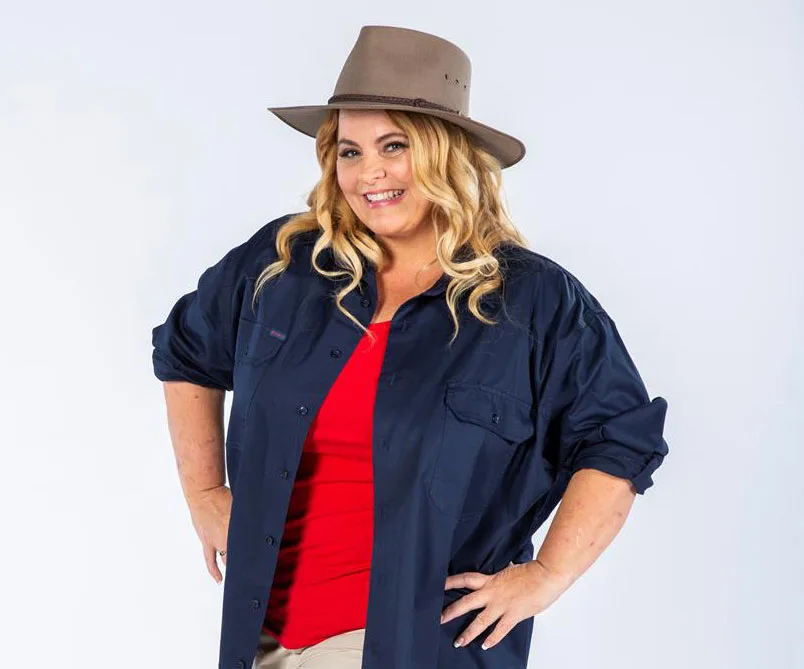 Smiling woman in blue shirt and red top, wearing a wide-brimmed hat, stands confidently with hands on hips.