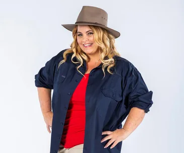 Smiling woman in blue shirt and red top, wearing a wide-brimmed hat, stands confidently with hands on hips.