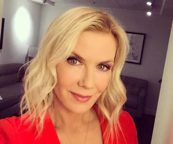 Blonde woman with wavy hair and red top smiling in a softly lit room with beige furniture in the background.
