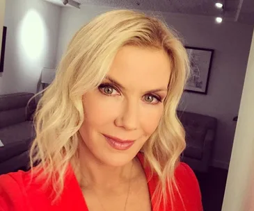 Blonde woman with wavy hair and red top smiling in a softly lit room with beige furniture in the background.