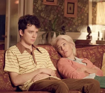 A teenage boy and his mother sit on a vintage couch in a cozy, retro-styled living room, with the mother looking at him.