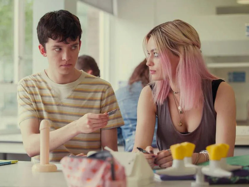 A young man and woman are in a classroom, with a model on the table; he is handing her a pen.