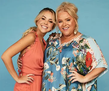Two women smiling, one in a coral dress and the other in a floral blue top, against a plain blue background.