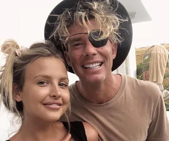 Two smiling people taking a selfie; one wears a black eyepatch and hat.