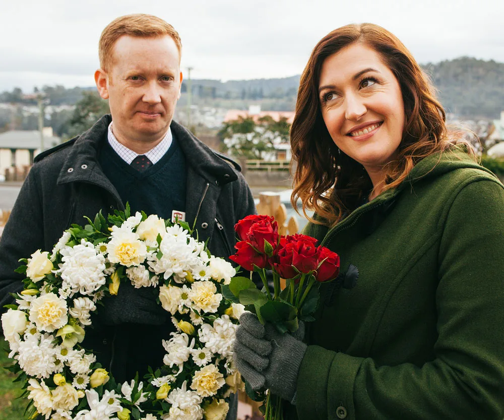 Man holding white flower wreath and woman holding red roses, smiling outdoors in overcast weather.