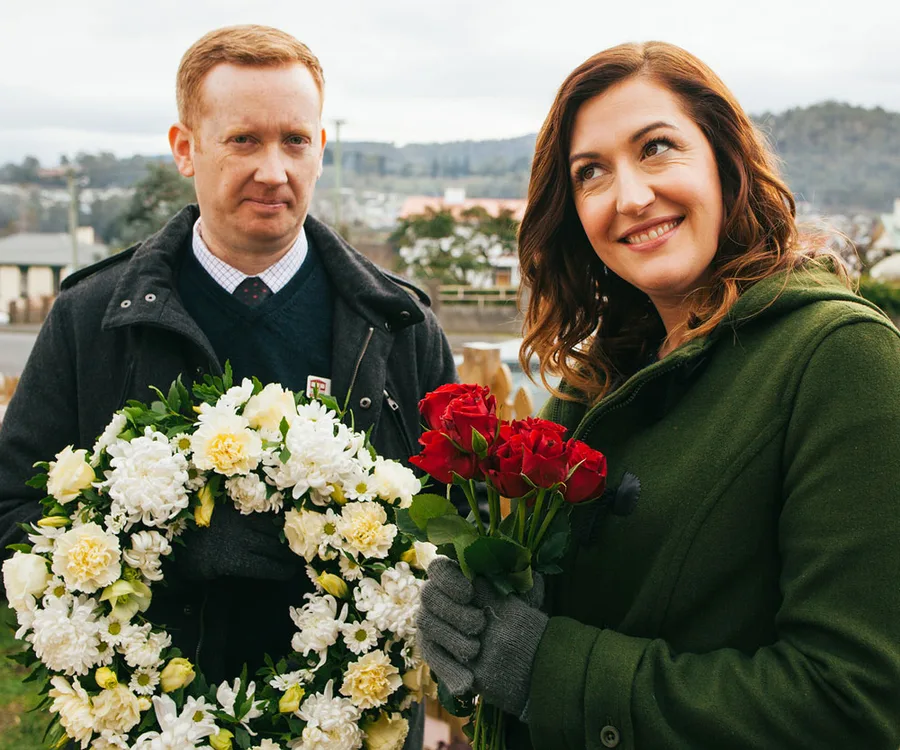 Man holding white flower wreath and woman holding red roses, smiling outdoors in overcast weather.
