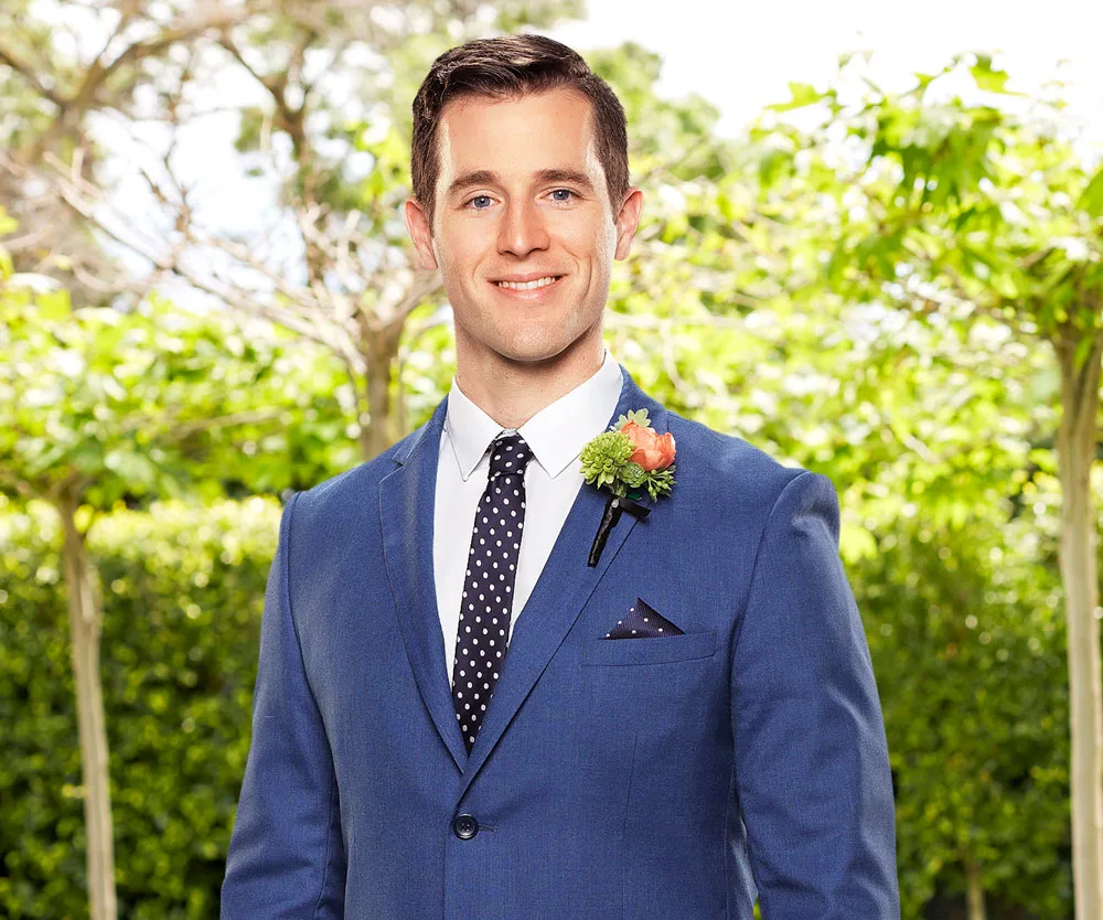 Man in blue suit with polka-dot tie and boutonniere, smiling in outdoor setting with greenery.