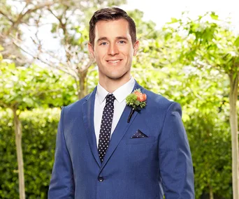 Man in blue suit with polka-dot tie and boutonniere, smiling in outdoor setting with greenery.