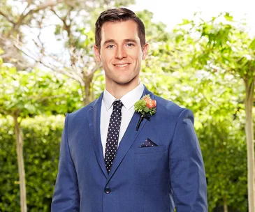 Man in blue suit with polka-dot tie and boutonniere, smiling in outdoor setting with greenery.