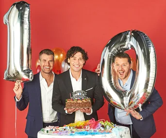 "Three men celebrate with birthday cake and large silver balloons forming the number 10 against a red background."