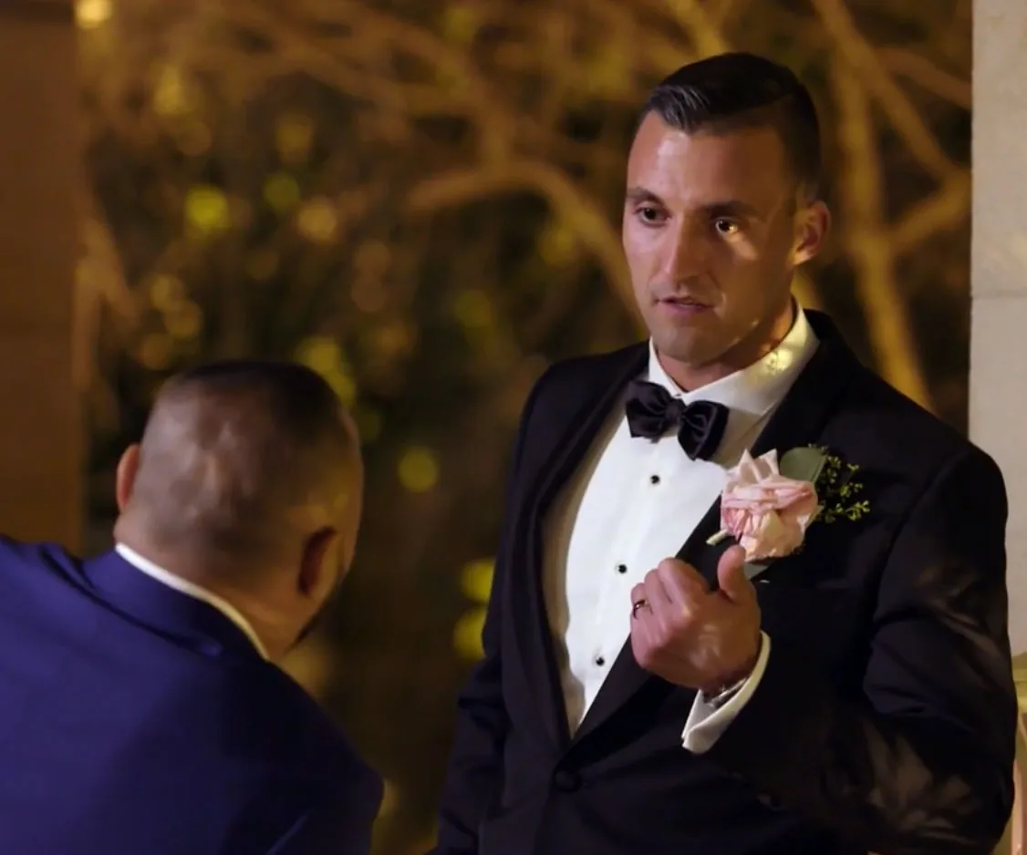A man in a tuxedo with a pink boutonnière gestures during a conversation in an outdoor setting at night.