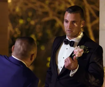 A man in a tuxedo with a pink boutonnière gestures during a conversation in an outdoor setting at night.