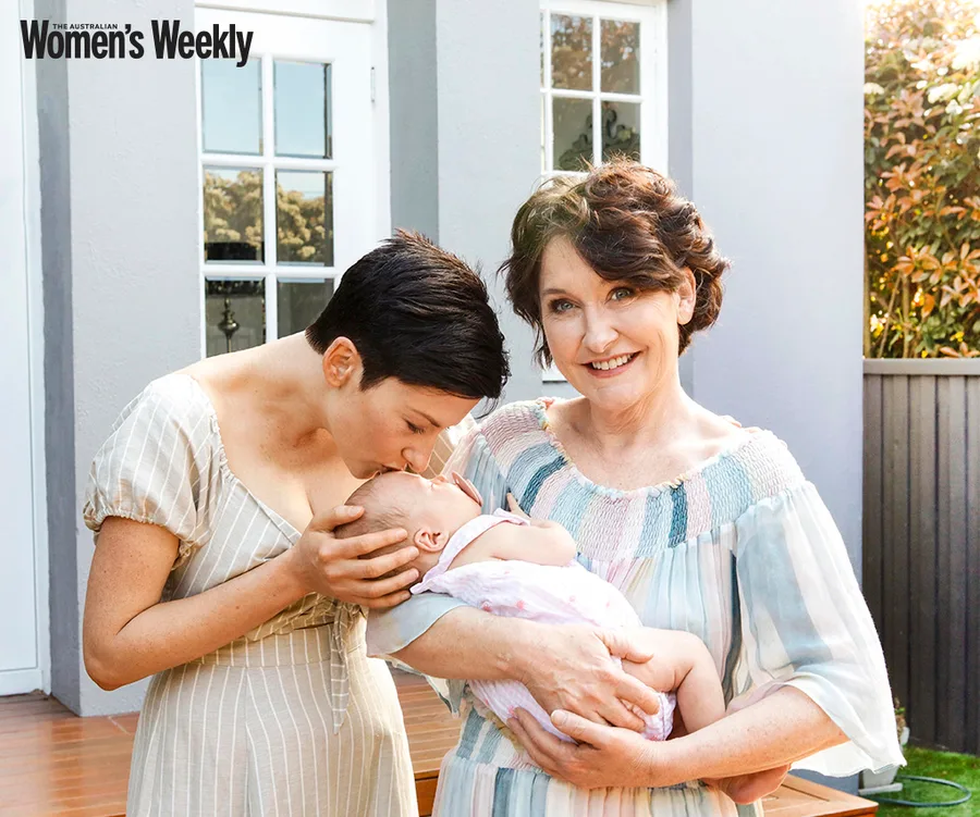 A woman smiles while holding a baby, and another woman kisses the baby's forehead outdoors.