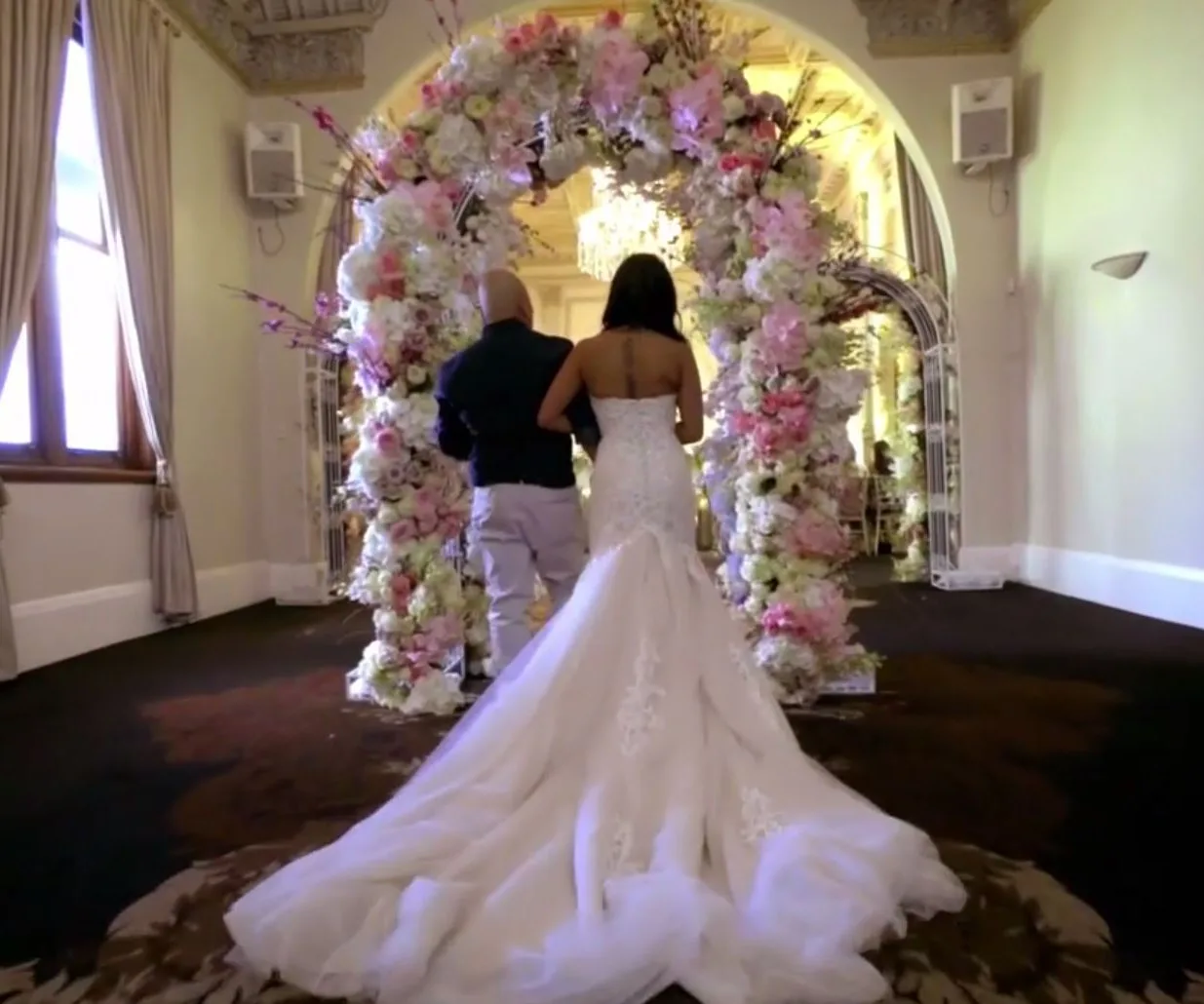 A bride in a white gown walks arm-in-arm with a man under a floral arch, in an elegant, softly-lit room.