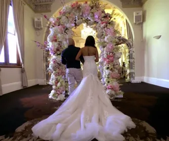 A bride in a white gown walks arm-in-arm with a man under a floral arch, in an elegant, softly-lit room.