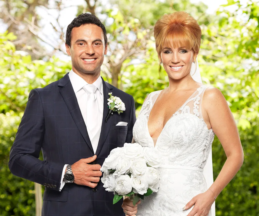 Bride in lace gown and groom in blue suit pose with bouquet in a garden setting.