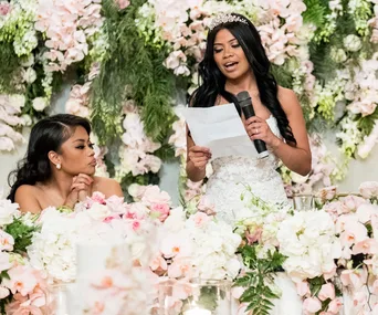 Two women at a floral-decorated wedding, one speaking into a microphone, the other listening attentively.
