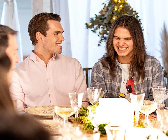 Two men are sitting at a dining table, smiling and laughing, surrounded by plates and glasses.