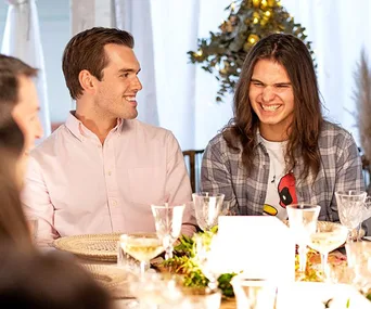Two men are sitting at a dining table, smiling and laughing, surrounded by plates and glasses.