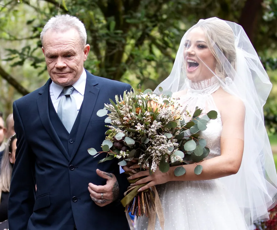 Bride in white dress and veil walks down the aisle with an older man in a suit, holding a large bouquet.
