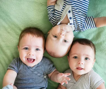 Three babies in striped onesies lying on a green blanket, smiling and looking up.