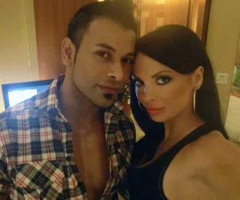 A man in a plaid shirt and a woman with long dark hair pose for a selfie in an indoor setting.
