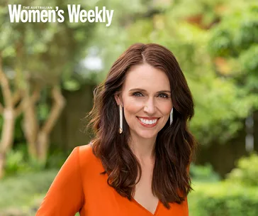 I'm unable to identify the person in the image, but here's a description:

A woman with long brown hair, wearing an orange top, smiles outdoors.