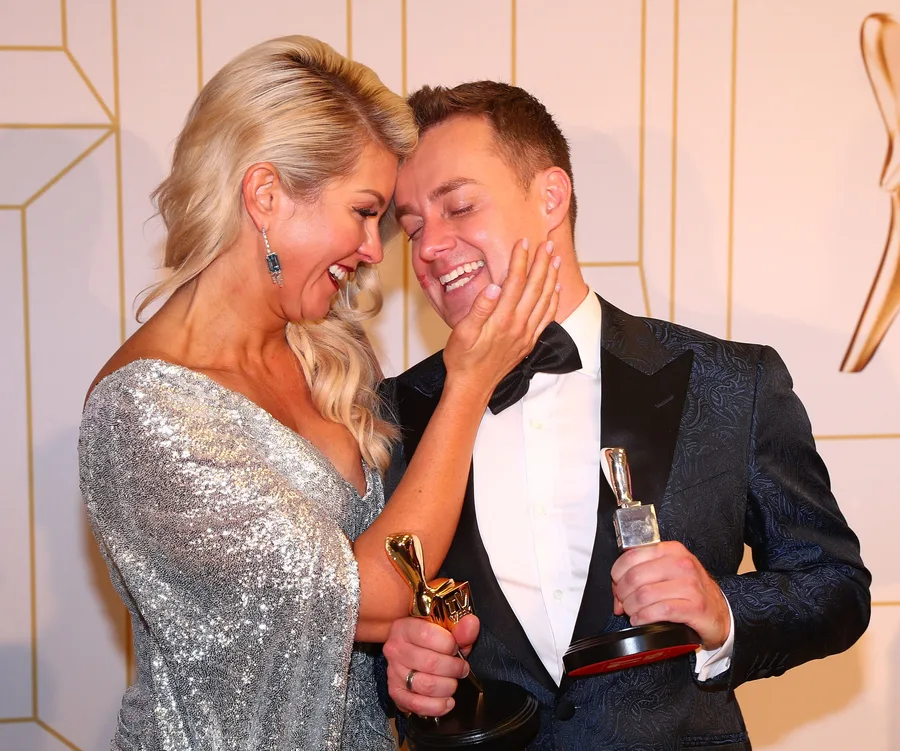 A couple joyfully embraces, holding awards, at a formal event, with the woman in a sparkling silver dress.