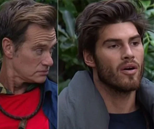 Two men in a jungle setting, one in red looking sideways, the other with a beard speaking, from "I'm a Celebrity."