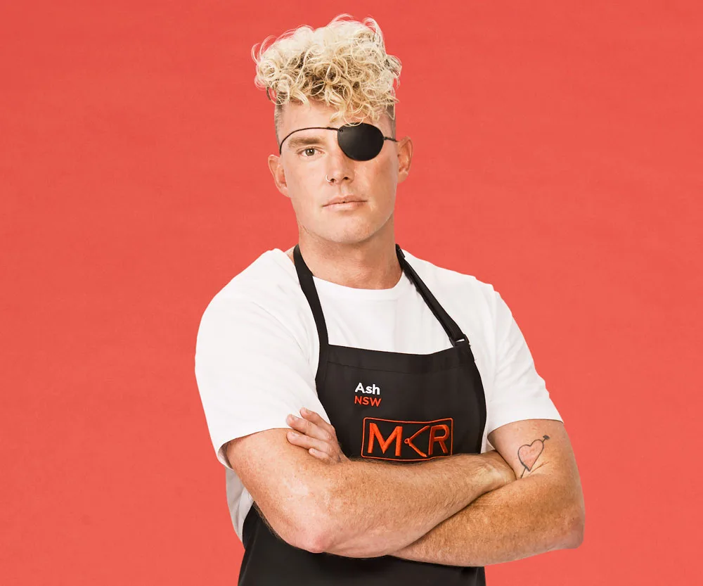 Man with blonde curly hair, wearing an eyepatch and "MKR" apron, arms crossed against a red background.