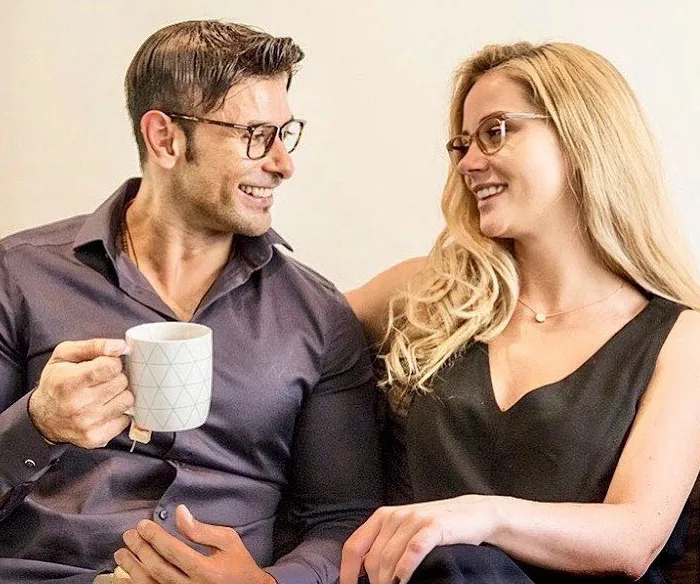 A smiling couple, both wearing glasses, sitting closely. One is holding a mug.