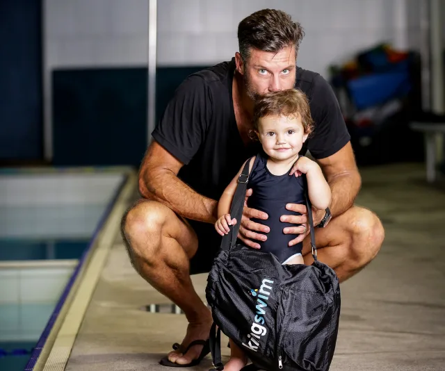Father and child in swimwear next to a pool, holding a bag labeled "Kingswim," ready for a swim lesson.