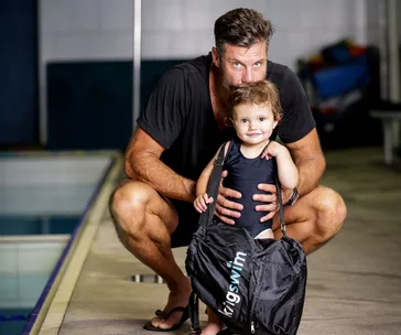 Father and child in swimwear next to a pool, holding a bag labeled "Kingswim," ready for a swim lesson.