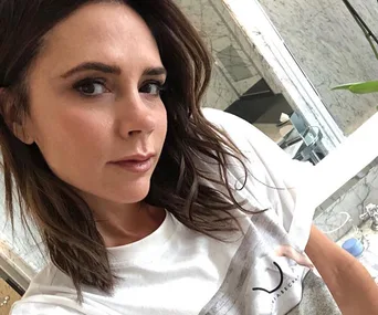 A woman with shoulder-length hair and a white shirt takes a selfie in a marble bathroom.
