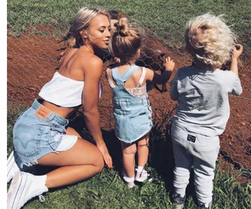 Woman and two children in casual clothes, interacting with a fenced area outdoors on a sunny day.