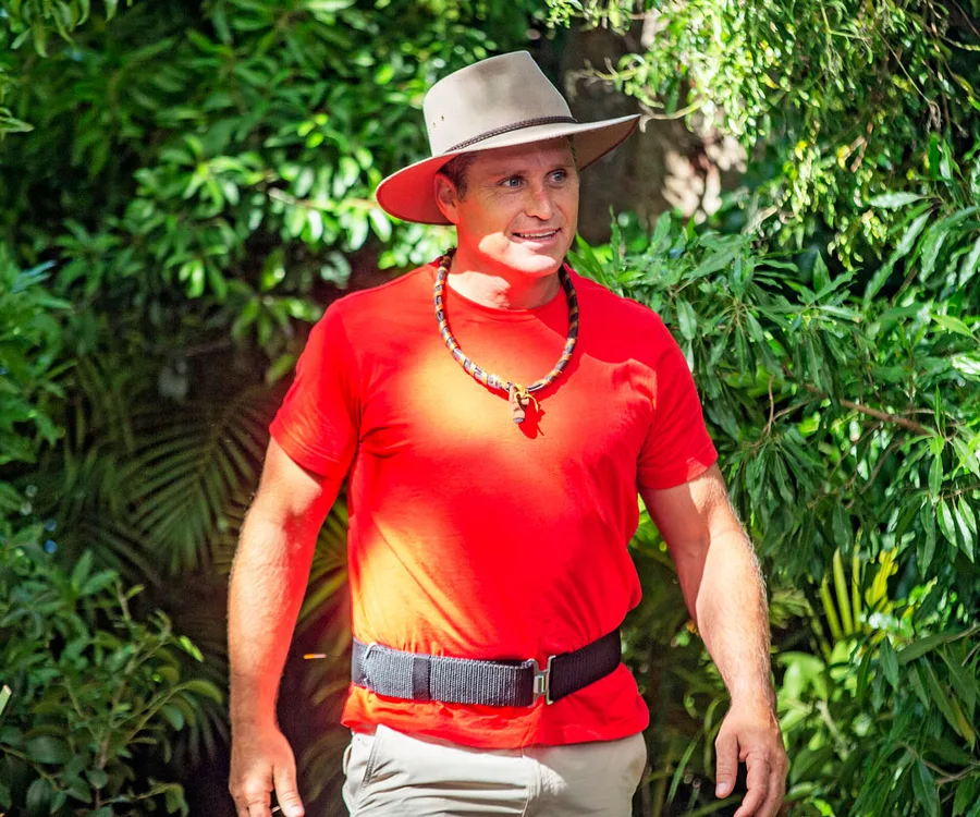 Man in a red shirt and beige hat walking through jungle setting.