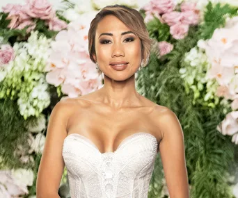 Ning Surasiang from Married at First Sight in a white dress, standing before a floral backdrop.