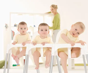Three babies in yellow onesies sit in high chairs, while a woman in the background tends to shelves.