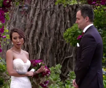 Wedding scene from “Married at First Sight” with bride in white and groom in dark suit by floral arch.