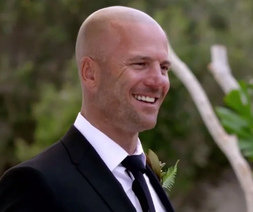 Man in a suit smiling outdoors with greenery in the background.