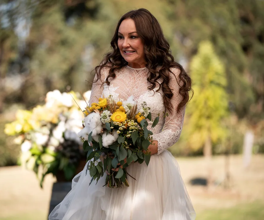 A woman in a lace wedding dress holds a bouquet of sunflowers and greenery outdoors.