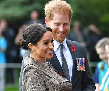 A smiling couple outdoors, the woman in checked attire and the man in a suit with medals, surrounded by greenery and people.