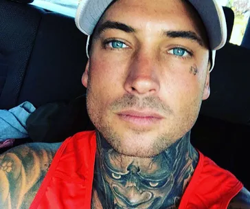 Young man with tattoos on neck and face, wearing white cap and red tank top, looking directly at camera in a car interior.