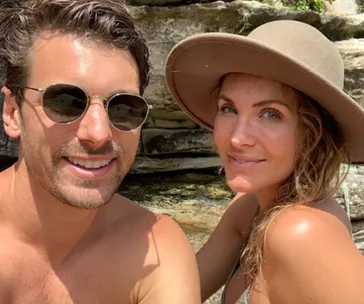 A couple smiling outdoors, man wearing sunglasses, woman in a hat, with rock formations in the background.