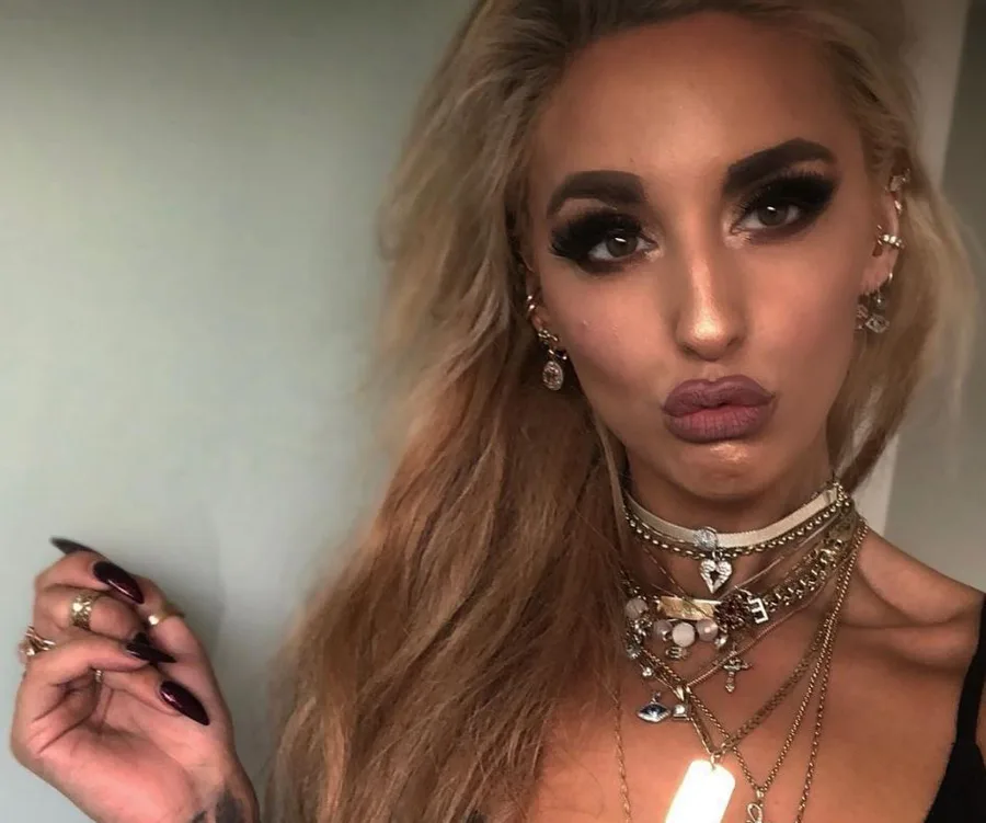 Woman with long hair, heavy eye makeup, pouting, wears layered necklaces and earrings.